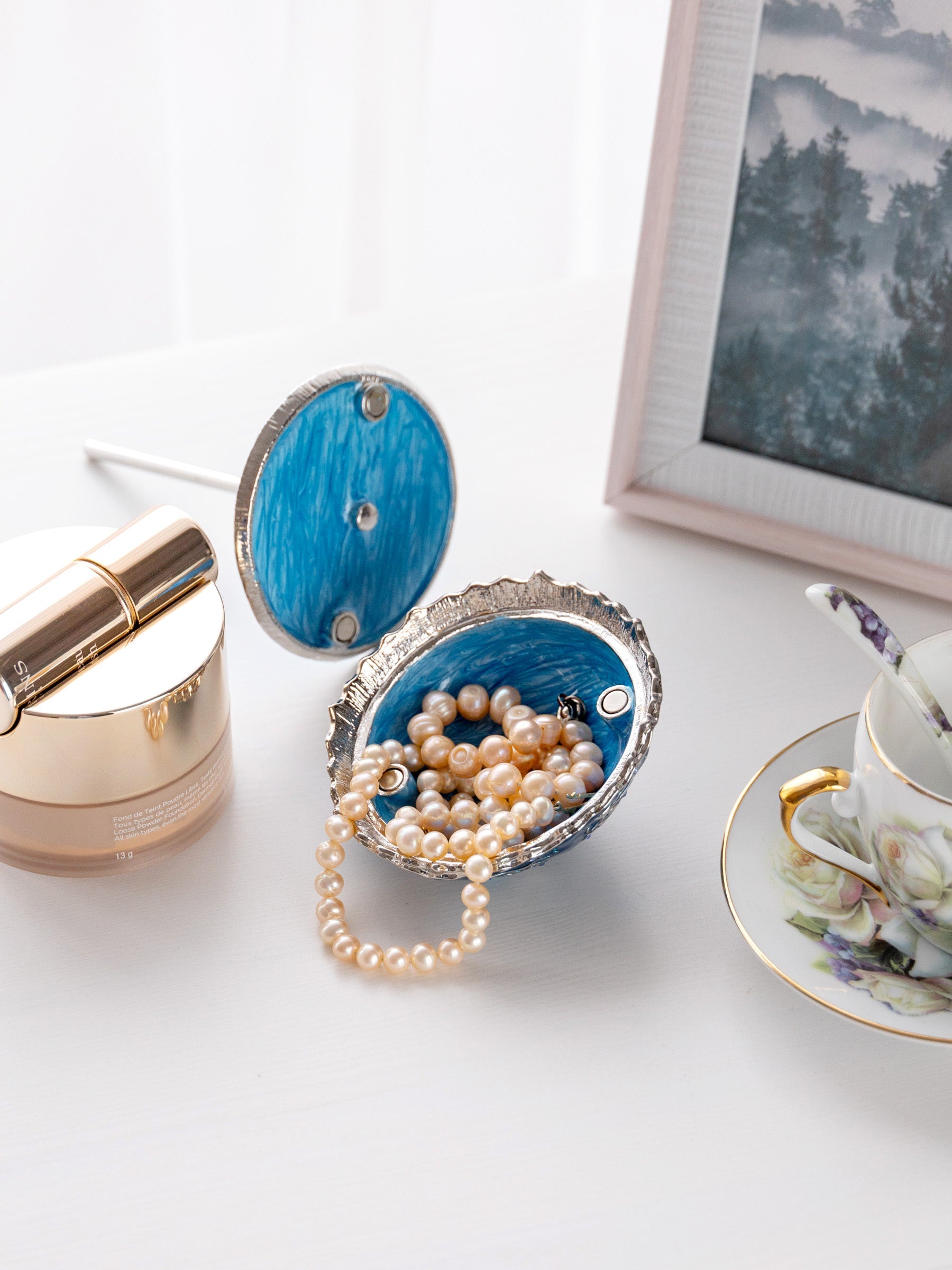 Blue jewelry box with pearls on a white surface next to a teacup and cosmetic product.