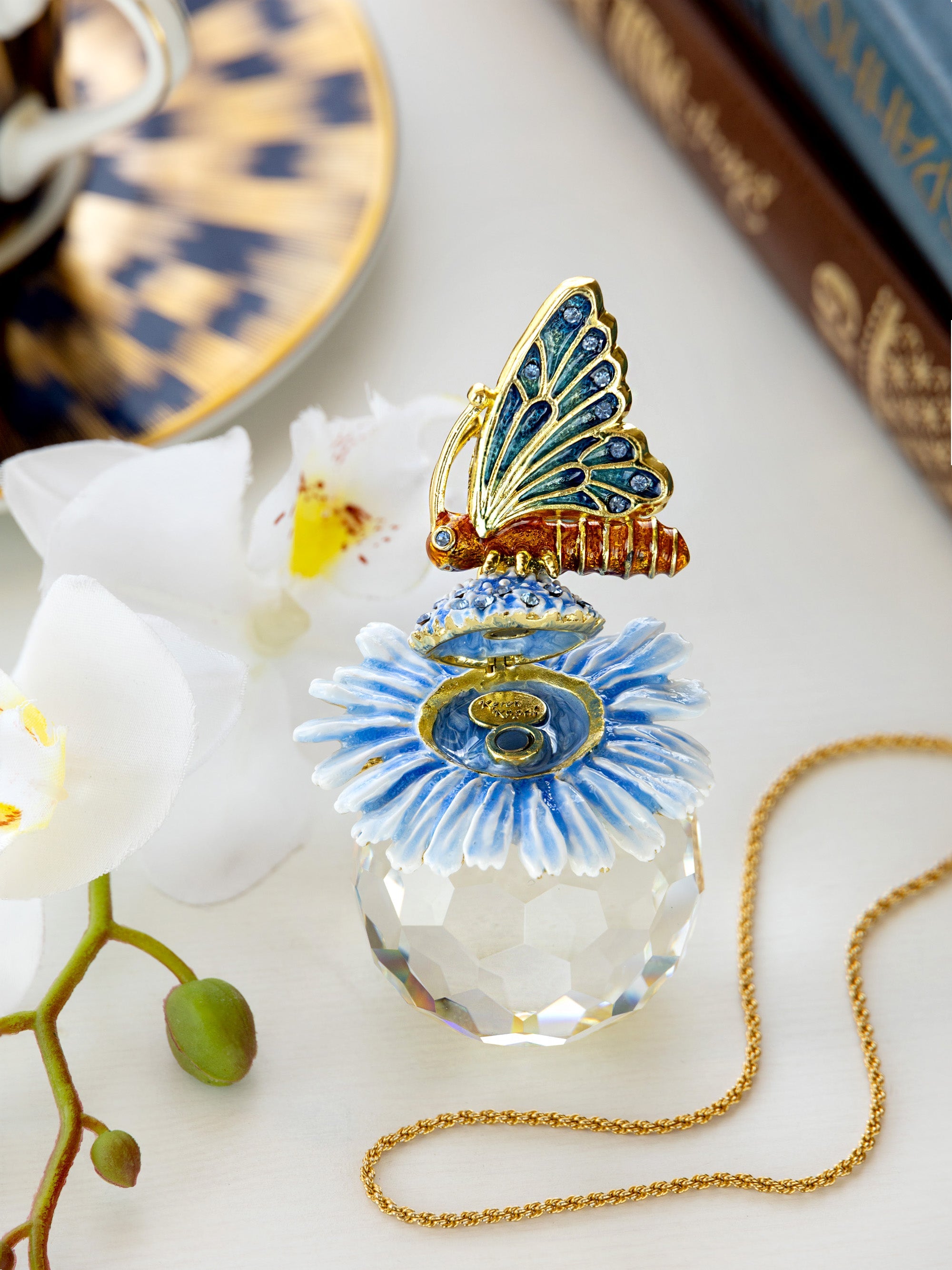 Decorative glass perfume bottle with butterfly top on a white surface with flowers and books in the background