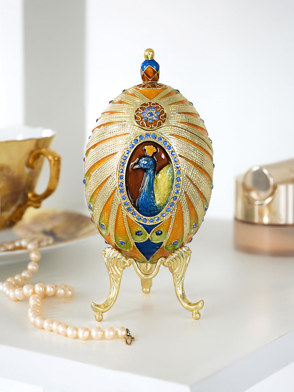 Decorative egg with peacock design on a stand, surrounded by pearls and tea set.