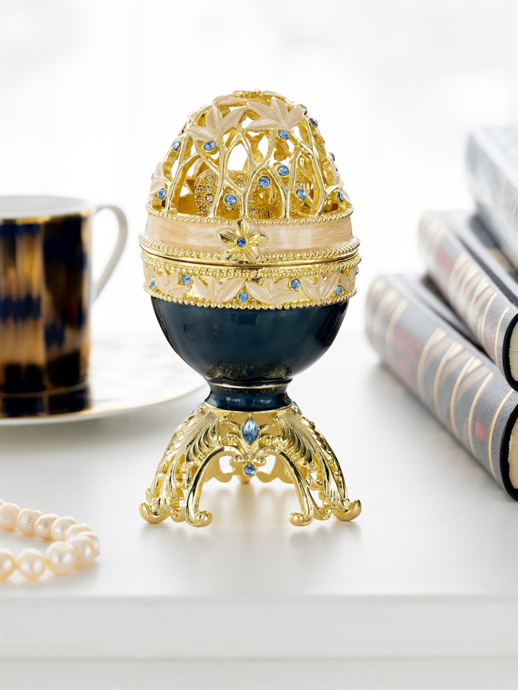 Decorative egg with gold and blue accents on a white surface with books and a cup in the background