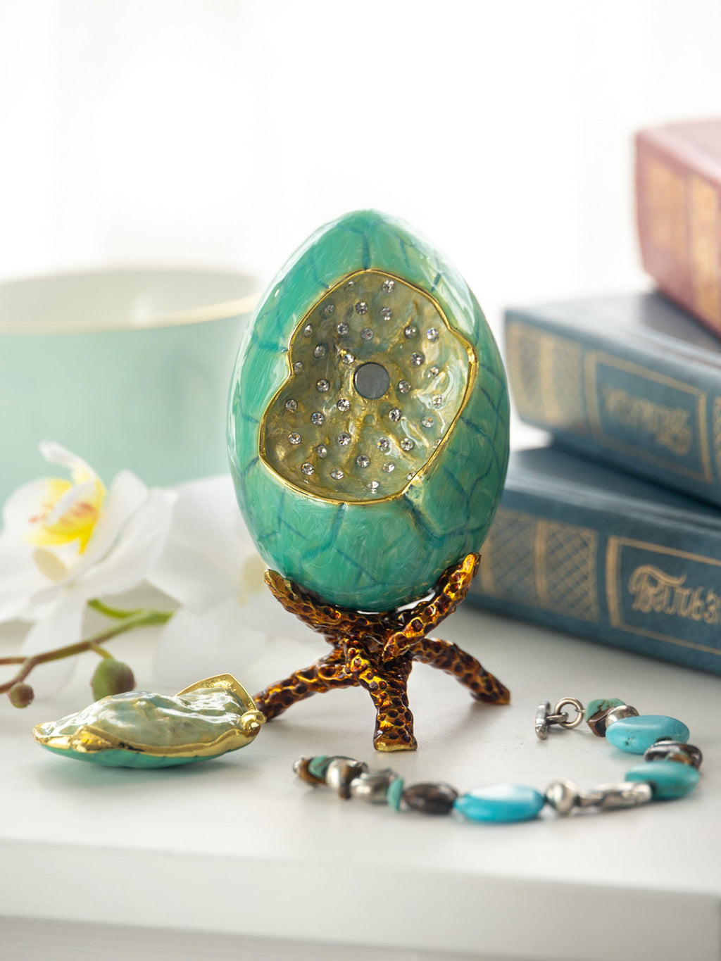 Decorative turquoise egg with gold accents on a stand, surrounded by books and flowers.